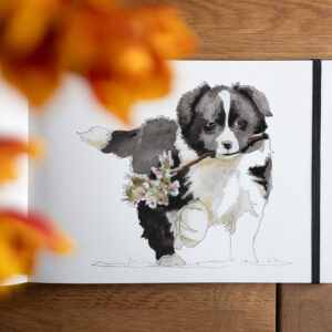 Aquarellierte Bleistiftzeichnung eines Border Collie Welpen, der einen blühenden Apfelzweig hält, in einem Skizzenbuch, das unter einem Strauß Tulpen auf einem braunen Holztisch liegt